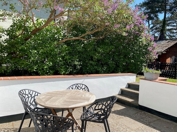 An outdoor patio with a round table and four chairs, surrounded by a low white wall and a flowering tree. - Strandhaus Clara: Large Terrace & Sea View at Timmendorfer Strand (Timmendorfer Strand)