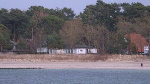 Cena costeira com uma praia de areia, um pequeno edifício branco e árvores densas ao fundo.