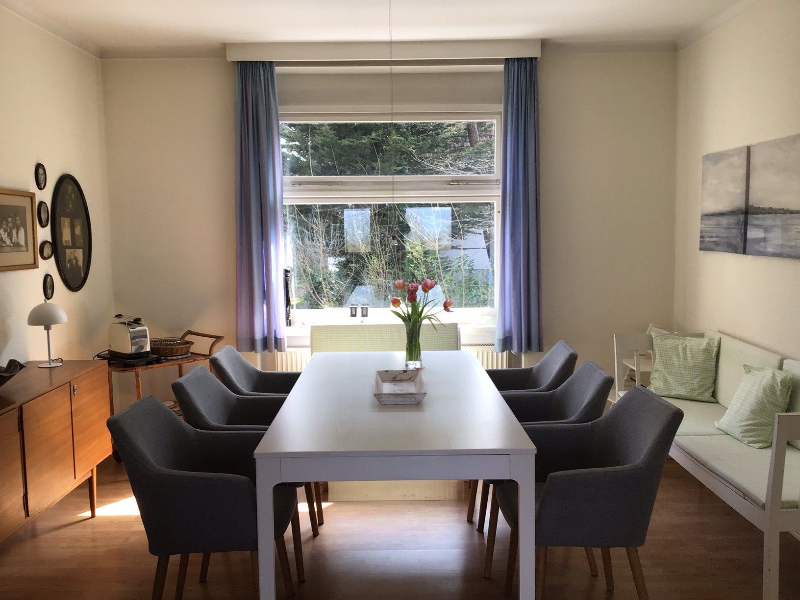 Un comedor con una mesa blanca, seis sillas, un jarrón con flores y una ventana con cortinas.