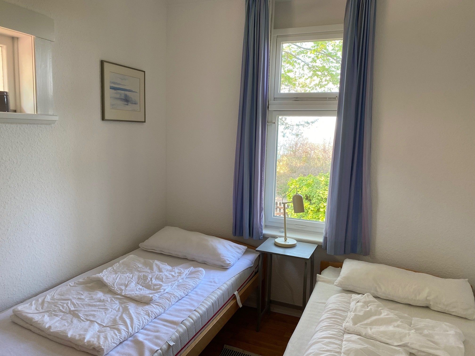 Ein schlichter Raum mit zwei Einzelbetten, einem kleinen Tisch mit Lampe und einem Fenster mit Vorhängen.