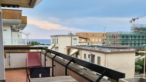 A balcony with a wooden table and chairs, a red chair, and a view of buildings and a construction site in the background. - Due Passi di Blu: Your own little oasis of relaxation just a few steps from the sea in Anzio (Anzio)