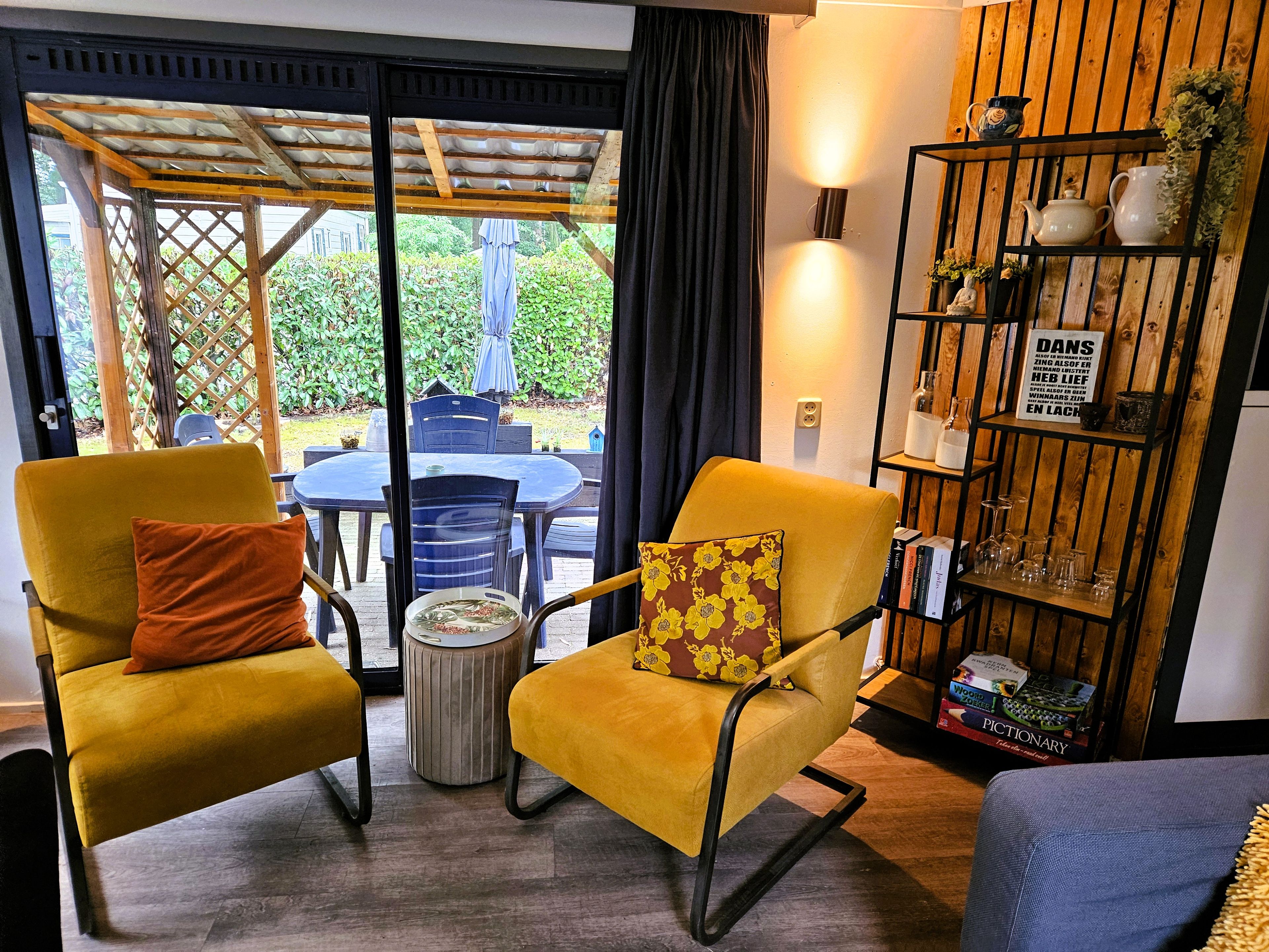 A room with two yellow armchairs, a small round table, a bookshelf with various items, and a view through a glass door to an outdoor area with a table and chairs.