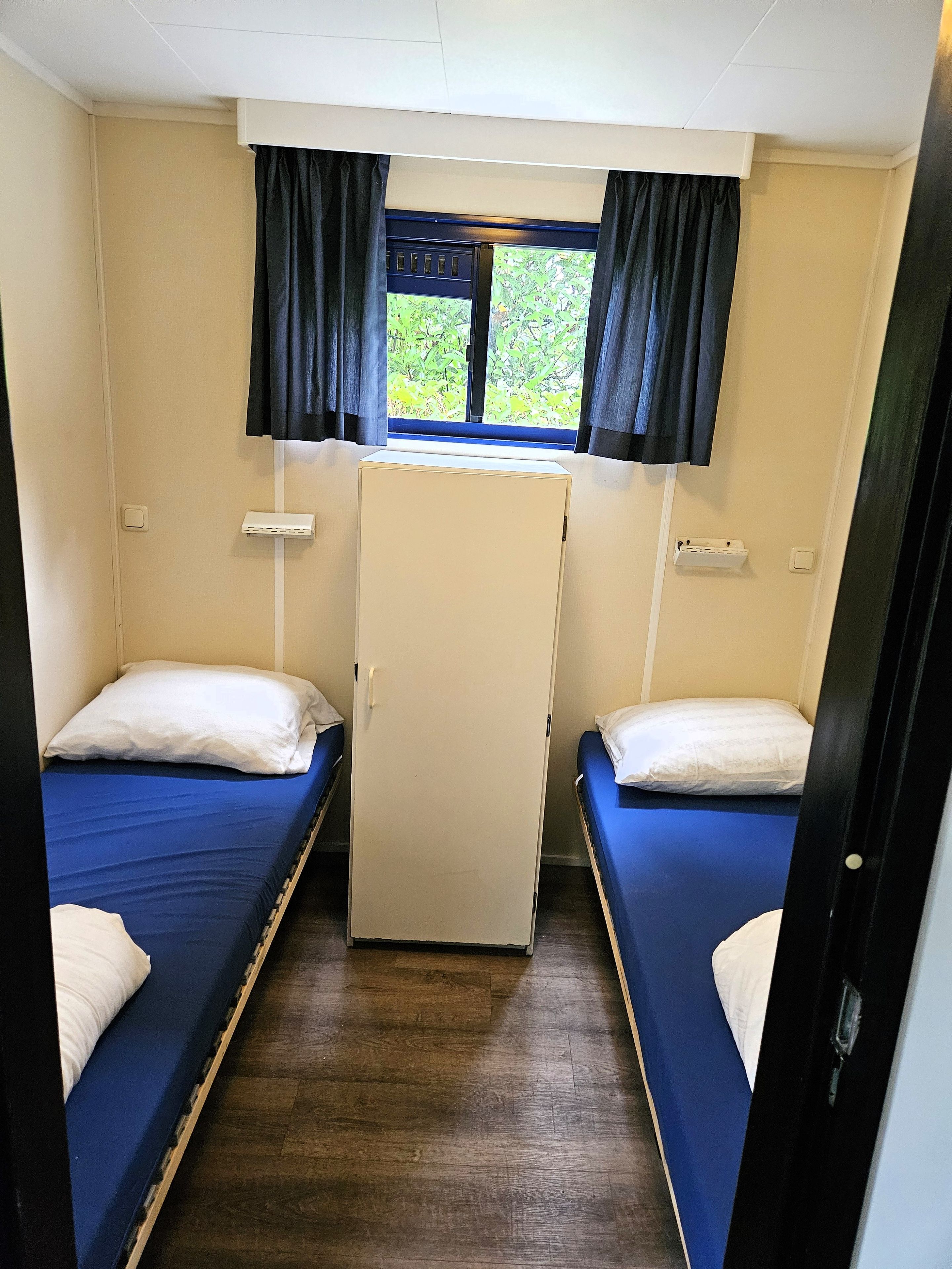 A small, compact room with two single beds, a small white cabinet, and a window with curtains.