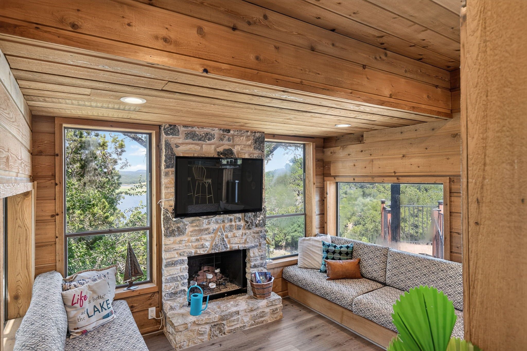 A cozy cabin interior with a stone fireplace, wooden walls, and large windows offering scenic views.