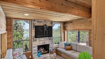 A cozy cabin interior with a stone fireplace, wooden walls, and large windows offering scenic views.