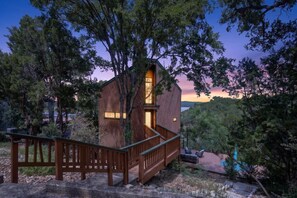 A wooden cabin with a staircase, surrounded by trees and a clear sky. - *New* Hill Country Treehouse Retreat | Pool, Hot Tub & Lake Travis Views ⛰️🌊 (Spicewood)