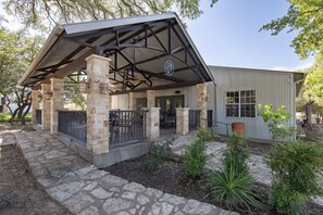 A covered outdoor seating area with stone pillars, a metal roof, and a building with a corrugated metal exterior. - *New* Hill Country Treehouse Retreat | Pool, Hot Tub & Lake Travis Views ⛰️🌊 (Spicewood)