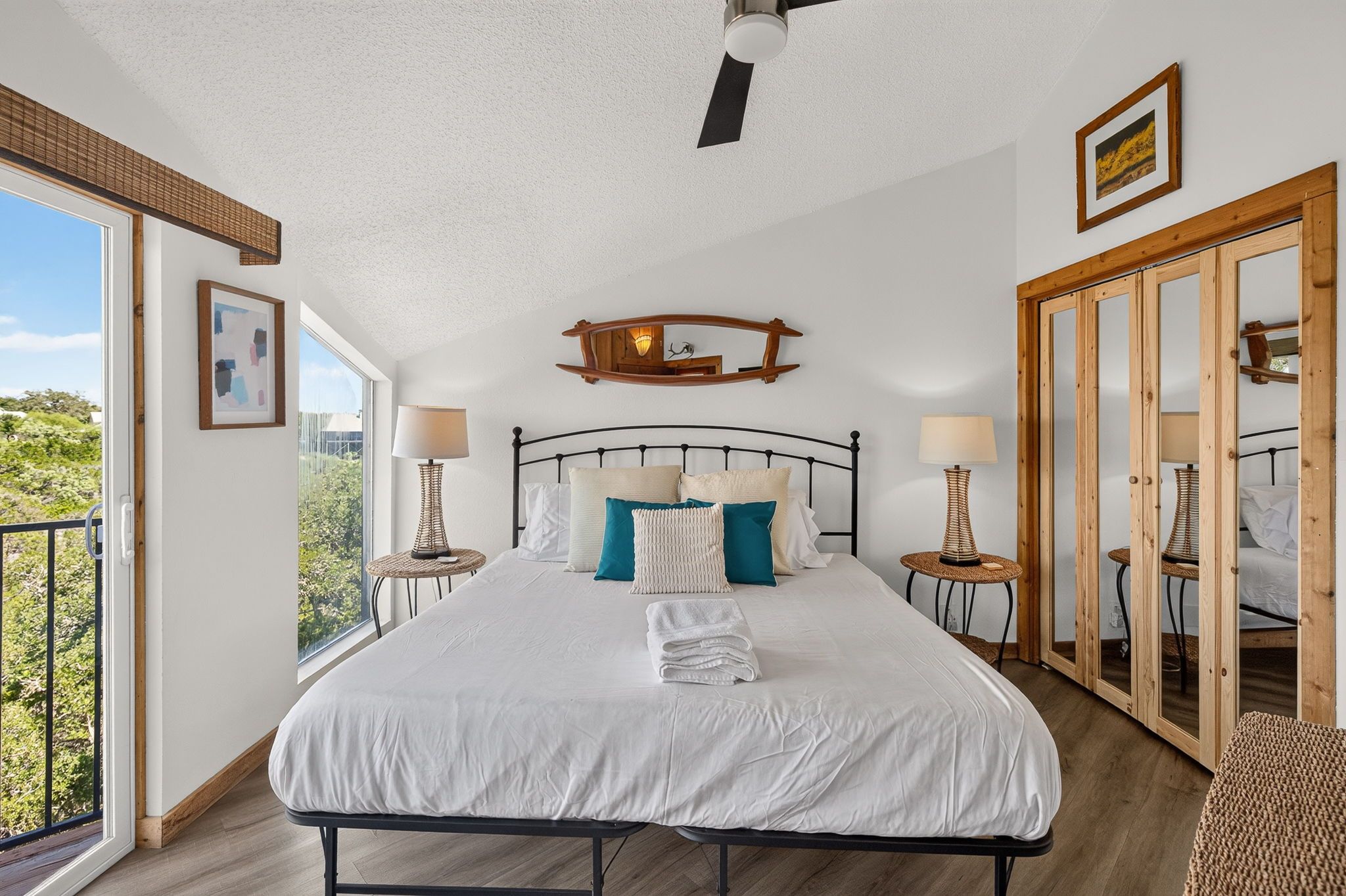 A bedroom with a large bed, two bedside tables with lamps, a ceiling fan, a wooden-framed mirror, and a view of greenery outside.