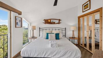 A bedroom with a large bed, two bedside tables with lamps, a ceiling fan, a wooden-framed mirror, and a view of greenery outside.
