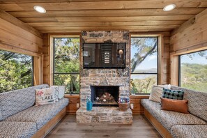 A cabin interior with a stone fireplace, wooden walls, and large windows offering scenic views.