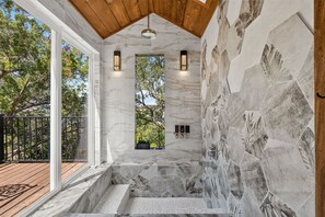 A bathroom with a stone wall, a wooden ceiling, and a view of trees outside. - *New* Hill Country Treehouse Retreat | Pool, Hot Tub & Lake Travis Views ⛰️🌊 (Spicewood)