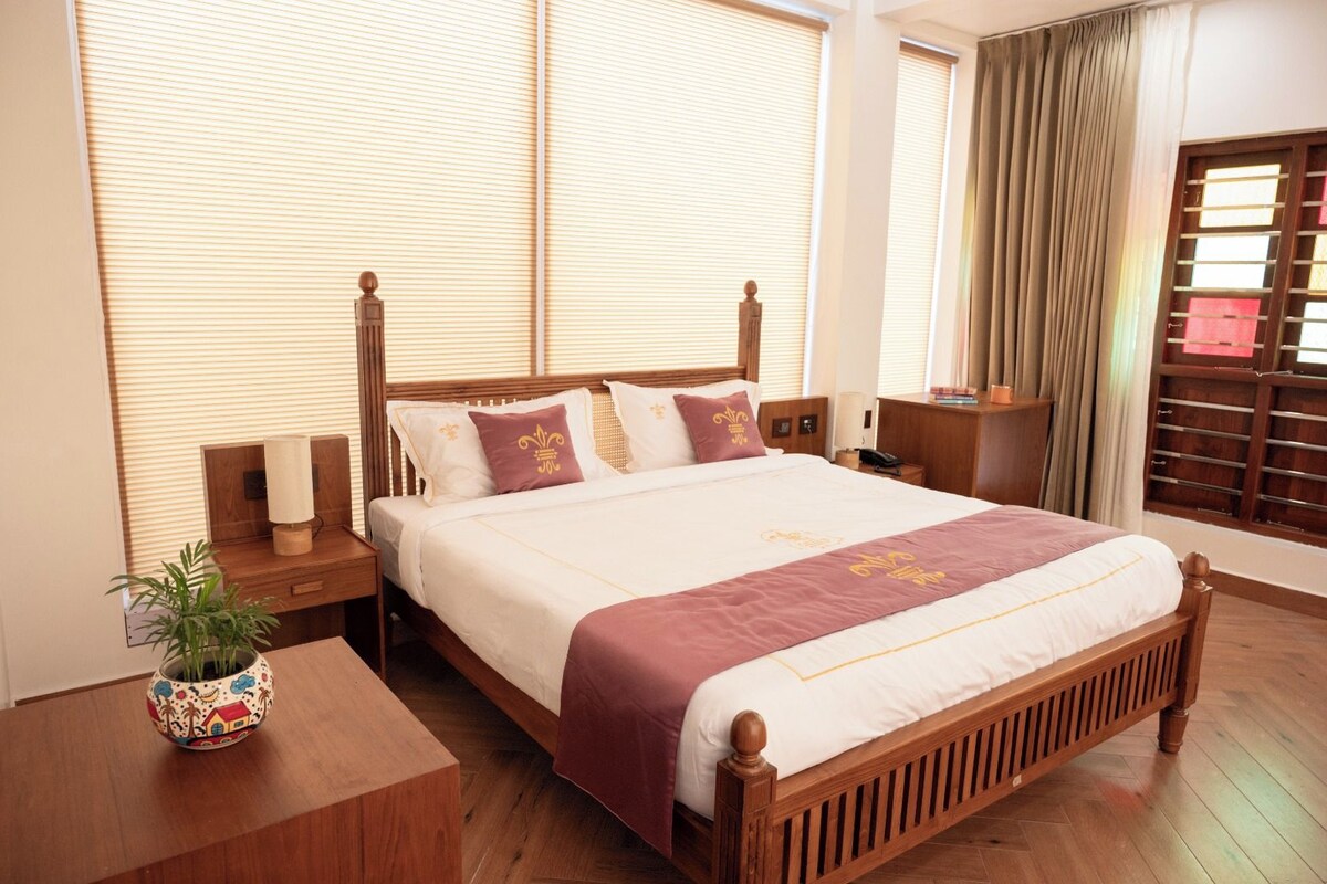 A hotel room with a large bed, wooden headboard, bedside tables, a nightstand, a bedside lamp, a decorative plant, and a window with blinds.