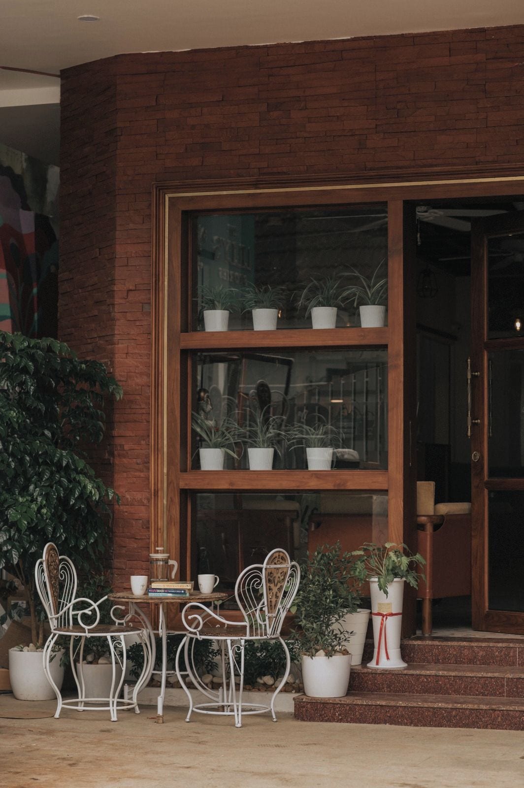 Un edificio de ladrillo con entrada por una puerta de cristal, un pequeño espacio al aire libre con sillas y mesa de metal, y plantas en macetas tanto dentro como fuera de la entrada.