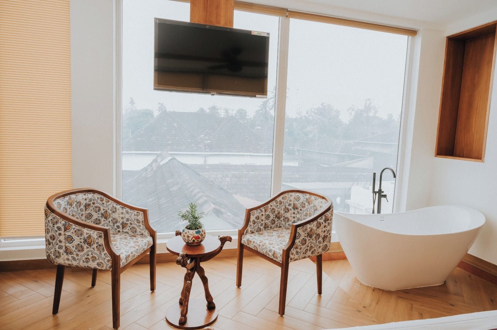 Un baño moderno con una bañera independiente, dos sillones estampados, una mesita con una planta y un ventanal con vistas a un paisaje urbano.