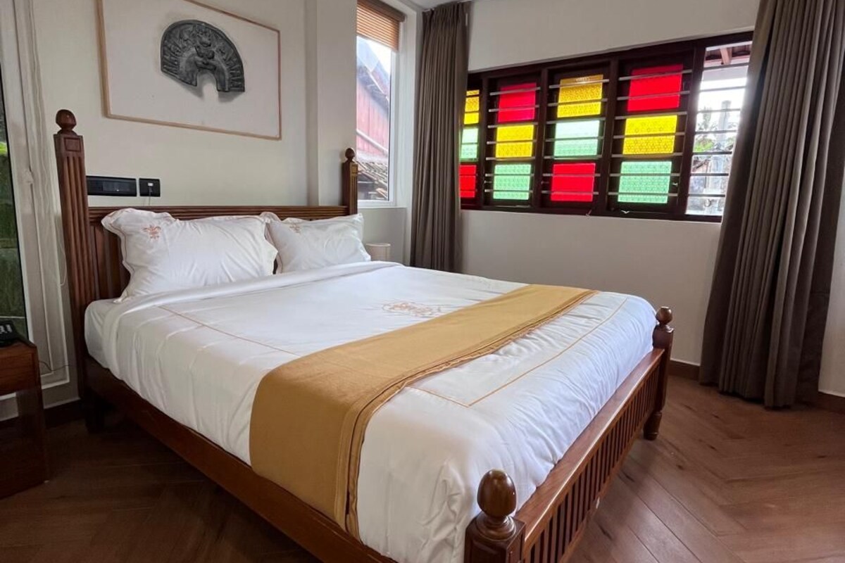 A hotel room with a wooden bed, a ceiling fan, a window with colorful stained glass, and a wooden floor.