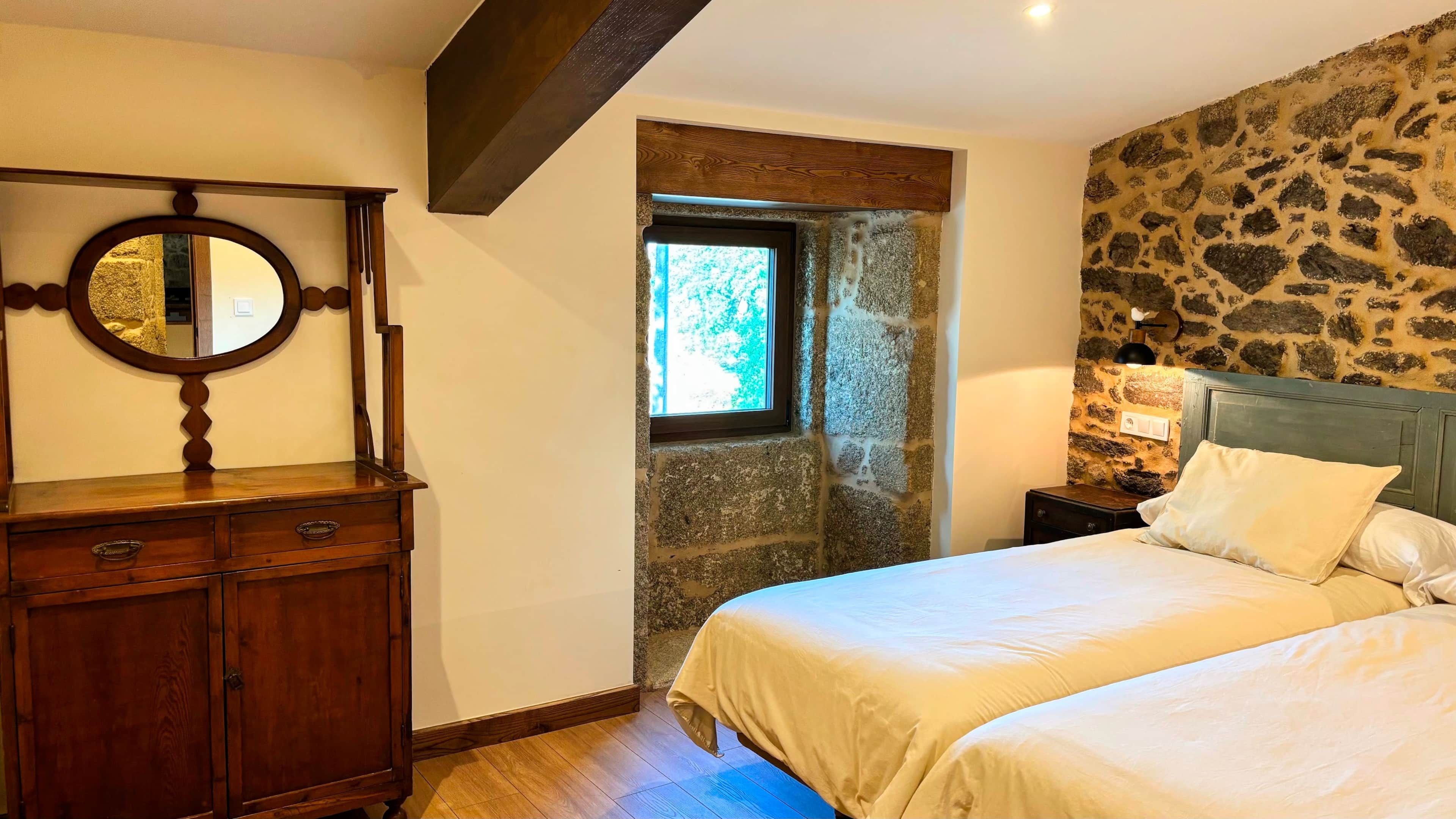 Un dormitorio con pared de piedra, cómoda de madera, cama con ropa blanca y mesita de noche con lámpara.