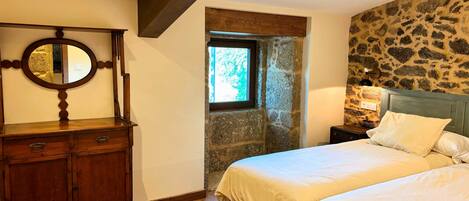 Un dormitorio con pared de piedra, cómoda de madera, cama con ropa blanca y mesita de noche con lámpara.