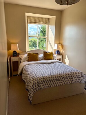 A bedroom with a large bed, two bedside tables with lamps, a window with a view of greenery, and a radiator. - The West Wing Sleeps 5 With Parking (Barnard Castle)