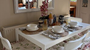 Et spiseområde med et hvidt bord dækket til fire personer, omgivet af hvide stole med mønstrede hynder.