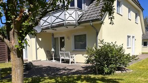 Uma casa amarela com varanda, um pequeno pátio com cadeiras e uma árvore ao primeiro plano.