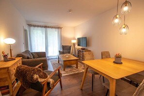 A cozy living room with a sofa, a coffee table, and a dining table with chairs. There is a television on the wall and a lamp on the side table.