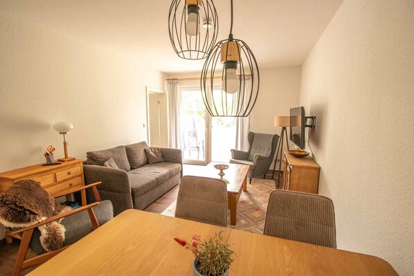 Ein modernes Wohnzimmer mit einer Couch, einem Esstisch mit Blumenvase, einem Holztisch und einem Fernseher.