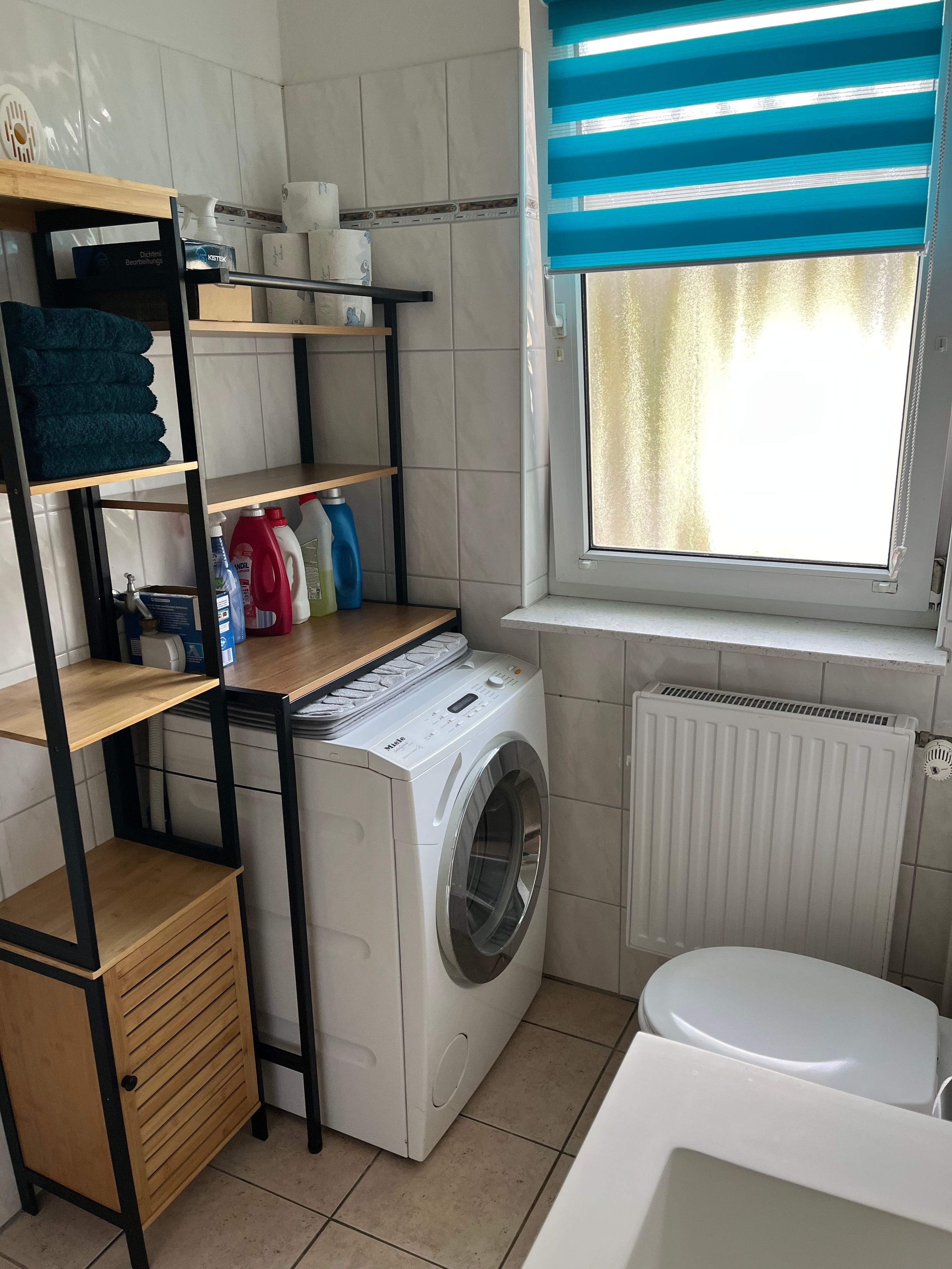 Un área de lavandería compacta con lavadora, repisas con productos de limpieza, un radiador y una ventana con persianas.