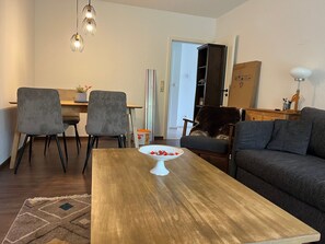 A living room with a wooden coffee table, two grey chairs, a dark sofa, and a dining area with a table and chairs.