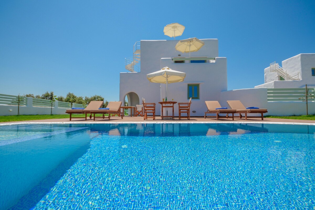 A swimming pool with lounge chairs and umbrellas, a white building with a staircase, and a clear blue sky.