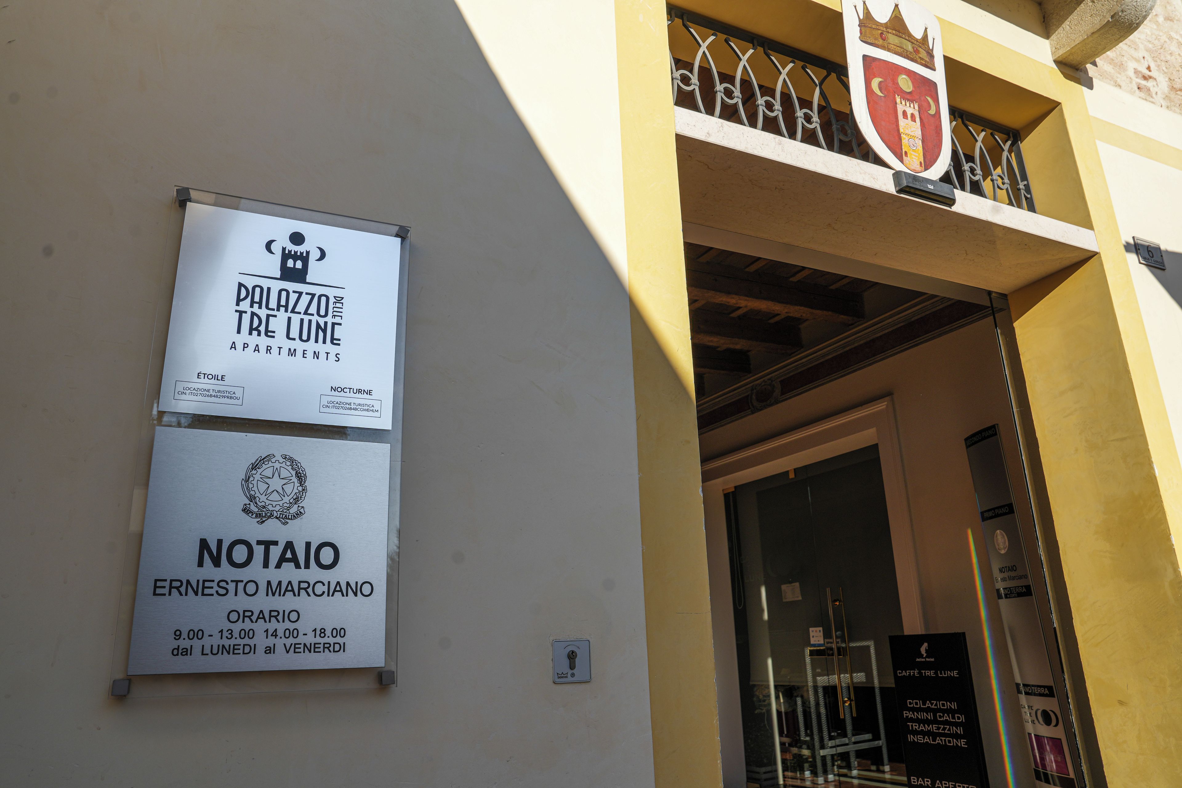 L’entrée d’un immeuble, avec une enseigne indiquant « Palazzo delle Tre Lune Apartments » et « Notaio Ernesto Marciano », ainsi que les heures d’ouverture.