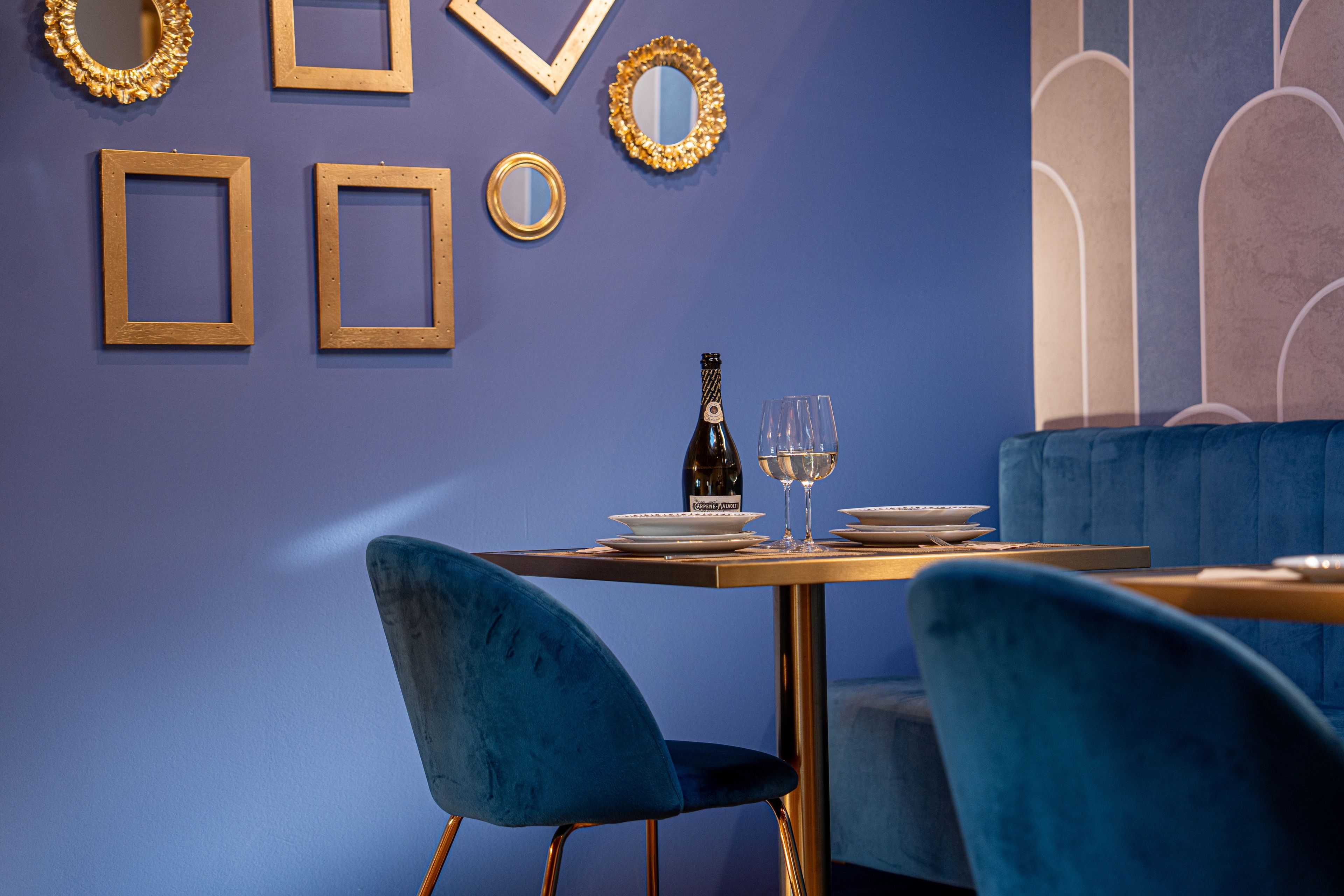A dining area with a table set for two, a bottle of wine, and glasses. There are framed pictures and mirrors on the wall.