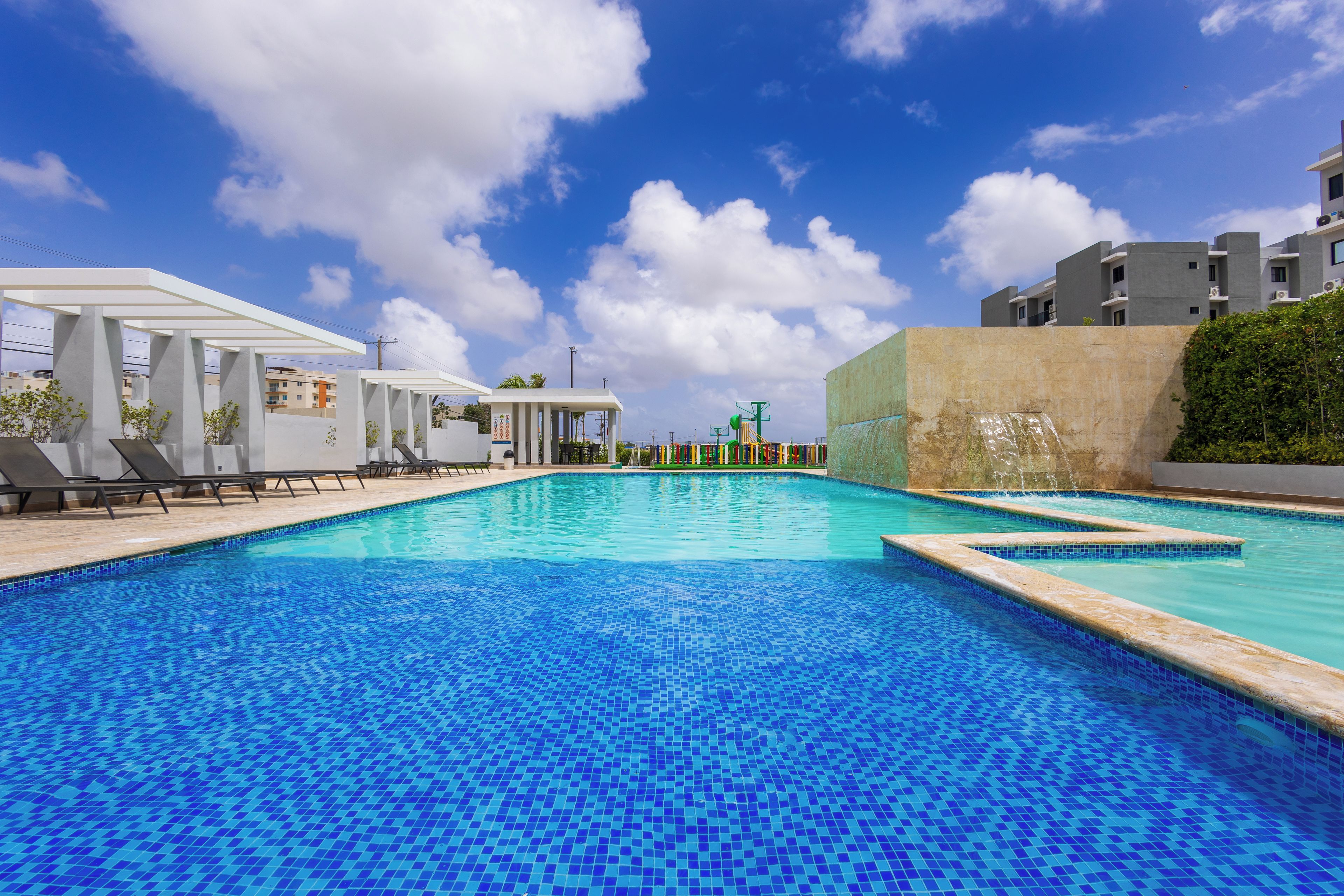 A swimming pool with a tiled blue surface, surrounded by lounge chairs and a covered area with a white roof.
