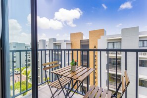 Ein Balkon mit einem Holztisch und Stühlen, einem Blumentopf und Blick auf moderne Gebäude unter blauem Himmel.
