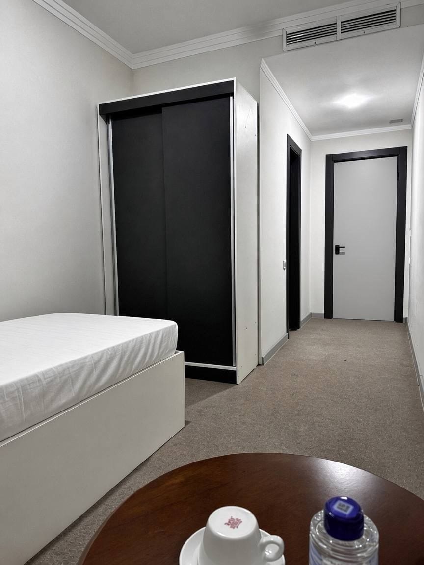 A hotel room with a bed, a wardrobe, a table with a cup and a bottle, and a door leading to another room.