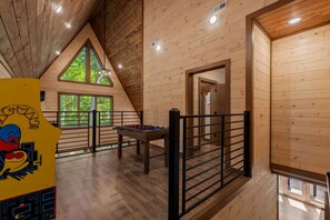 Une chambre spacieuse aménagée en loft, avec un plafond haut, des murs en bois, une table de baby-foot et de grandes fenêtres laissant entrer la lumière naturelle.