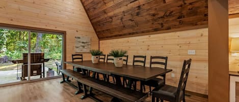Comedor de madera con una larga mesa y bancos, rodeado de sillas, con vista a espacios verdes.