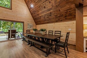 Un coin repas en bois, avec une longue table et des bancs, entouré de chaises, et offrant une vue sur la verdure à l’extérieur.