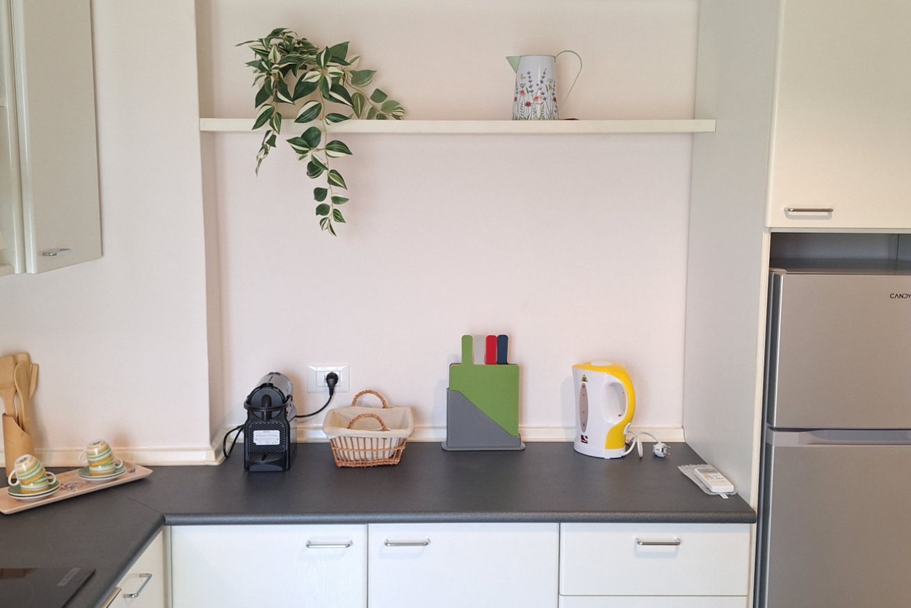 Een keuken met een zwart aanrecht, witte kasten, een plant, een waterkoker en een mand.