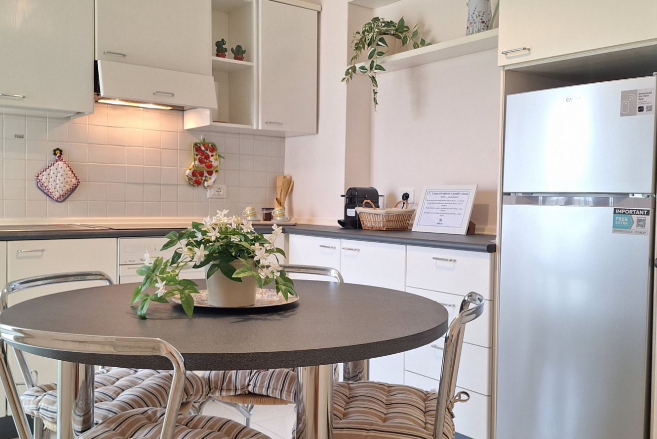 A modern kitchen with white cabinetry, a stainless steel refrigerator, a round dining table with chairs, and decorative plants.
