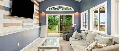 Una sala de estar con un televisor de pantalla plana montado en una pared de madera, una mesa de centro de vidrio, un sofá gris y vistas a una terraza de madera a través de una puerta de cristal.