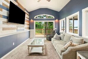Ein Wohnzimmer mit Flachbildfernseher an einer Holzwand, einem Glastisch, einem grauen Sofa und Blick auf eine Holzterrasse durch eine Glastür.