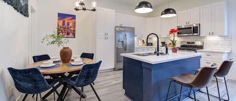 A modern kitchen with white cabinets, a central island with bar stools, and a dining area with blue chairs.