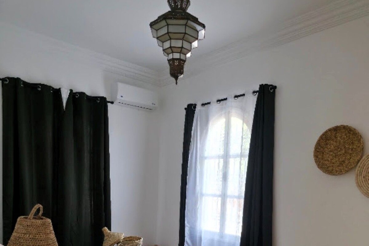 A room with a bed, a lamp, a window with curtains, and a woven mat on the floor.