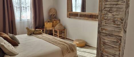 Un dormitorio con cama, espejo, armario y ventana con cortinas.
