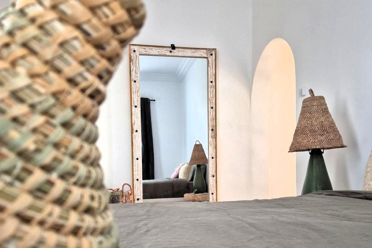 Una habitació amb un llit, una cadira de veta, una làmpada i un mirall que reflecteix una porta.