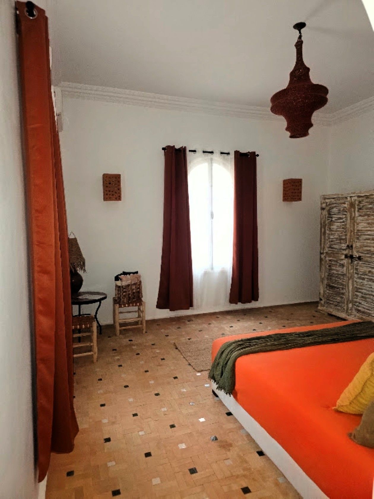 Una habitación con cama, una mesa, sillas, un ventanal con cortinas y una lámpara colgante.