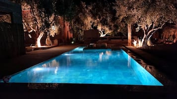Una piscina rectangular de noche, rodeada de árboles y una estructura de piedra.