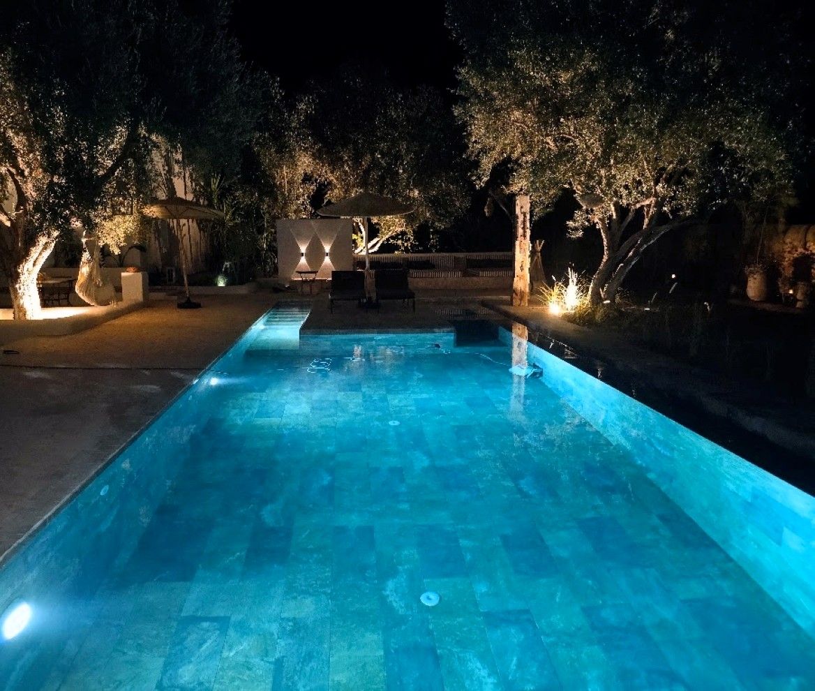 Una piscina nocturna con árboles alrededor y un pabellón.