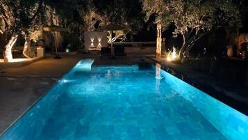Una piscina nocturna con árboles alrededor y un pabellón.