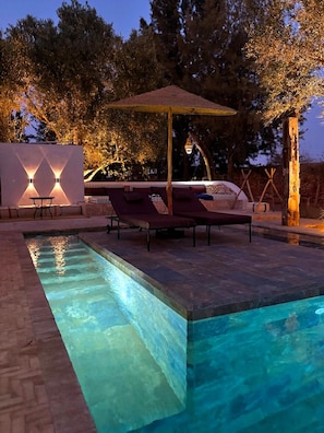 A swimming pool with lounge chairs and a pergola, surrounded by trees and a stone wall.